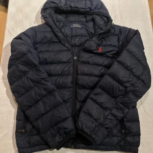Mörkblå dunjacka Polo Ralph Lauren - Snygg mörkblå dunjacka från Polo Ralph Lauren med huva och dragkedja. Jackan har det klassiska röda Polo-logot broderat på bröstet och två fickor med dragkedja. Quiltad design och normal passform, perfekt för kyliga dagar. OBS HAR ETT PYTTE LITET HÅL I ÄRMEN MEN DEN SYNS EJ MED BLÅTTA ÖGAT