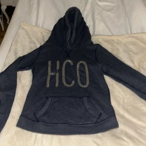 Mörkblå hoodie från Hollister XS - Mysig mörkblå hoodie från Hollister med stor HCO-logga i silver framtill. Tröjan har huva med snörning, känguruficka och långa ärmar. Perfekt för chill dagar och enkel att matcha med jeans eller leggings.