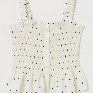 Vit prickig linnetopp med volang - Supersöt vit linnetopp med svarta prickar, smockad överdel och volang nertill. Breda axelband med rynkade detaljer och knäppning framtill med små knappar. 