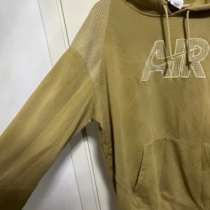 Beige grön Nike AIR hoodie  - Säljer en beige Nike AIR hoodie i storlek XL junior lmed meshdetaljer på axlarna och stor AIR-logga framtill. Tröjan har huva med snörning, känguruficka och långa ärmar. Perfekt för dig som gillar sportig och avslappnad stil. Pris kan diskuteras