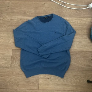 Blå stickad tröja från Polo Ralph Lauren - Snygg blå stickad tröja från Polo Ralph Lauren med rund hals och diskret broderad logga på bröstet. Tröjan har långärmad passform och ribbade muddar vid ärmslut och nederkant. Perfekt för dig som gillar klassisk stil med en modern touch.