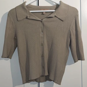 Beige ribbad blus med knappar - Säljer en beige ribbad blus från Uniqlo med klassisk krage och knäppning framtill. Blusen har trekvartsärm och är tillverkad i mjukt stickat material som ger en skön och avslappnad känsla. Perfekt för dig som gillar stilrena och enkla plagg.