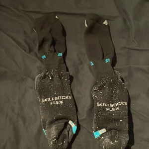 Svarta Skillsocks Flex träningsstrumpor - Svarta träningsstrumpor från Skillsocks Flex med vita och turkosa detaljer. Undersidan har vita prickar för extra grepp och texten 'SKILLSOCKS FLEX' på ovansidan. Perfekta för sport och aktivitet, med en normal passform och syntetmaterial.