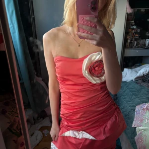 Rosa axelbandslös kortklänning Creativity - Säljer en superfin axelbandslös kortklänning i stark rosa från Creativity. Klänningen har draperad passform, volangdetaljer och en stor vit och rosa blomapplikation med pärlor framtill. Materialet är glansigt och känns lyxigt, perfekt för dig som vill sticka ut.