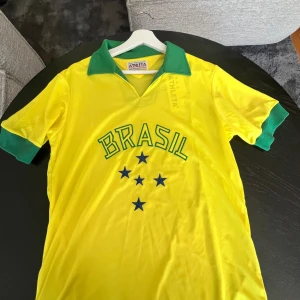 Brasilien fotbollströja Athleta gul - Retro Brasilien fotbollströja från Athleta i starkt gul polyester med gröna detaljer på krage och ärmslut. Klassisk design med 'BRASIL' och fem blå stjärnor tryckta på bröstet. Perfekt för dig som älskar vintage och fotbollshistoria.