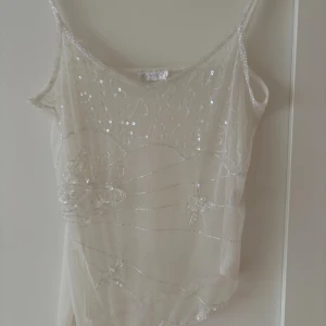Vit paljettprydd linnetopp - Säljer en vit linnetopp med tunna axelband och transparent mesh. Toppen är dekorerad med paljetter och broderade fjärilar samt blommor, vilket ger en glittrig och romantisk känsla. Perfekt för sommarens alla tillfällen.