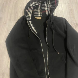 Svart Burberry hoodie med dragkedja som är sönder  - Dragkedjan är sönder men de går o fixas nästa överallt för 20kr storlek xs /s Svart hoodie från Burberry med klassiskt rutigt foder i huvan och silverfärgad dragkedja. Tröjan har snörning vid huvan och är tillverkad i mjuk bomull med lite elastan för extra komfort. Perfekt för dig som gillar stilrena och lyxiga streetwear-plagg.