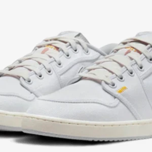 Nike Jordan 1 Low ajko - Nike Jordan 1 Low ajko i fräsch vit canvas med klassisk Swoosh i ljusgrå mocka. Skorna har snörning, platt sula och diskreta gula detaljer vid hälen. Perfekt för dig som gillar clean och stilren streetwear.