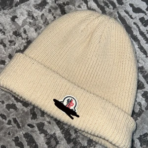 Beige stickad mössa  - Snygg beige stickad mössa med klassisk uppvikt kant och broderad logotyp framtill. Perfekt för att hålla värmen och samtidigt se clean ut. Passar dig som gillar stilrena accessoarer med exklusiv känsla.  !Inga taggar!