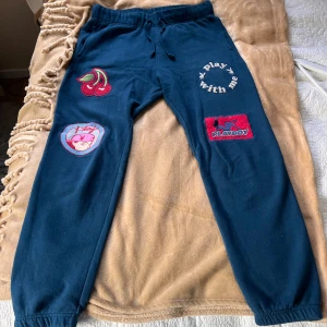 Blå Playboy by Pacsun mjukisbyxor M - Mörkblå mjukisbyxor från Playboy by Pacsun med broderade patches, körsbär, texten 'play with me' och flera Playboy-loggor. Byxorna har snörning i midjan, resår vid benslut och är gjorda i mjukt bomullsmaterial. Perfekta för en chill och trendig look.