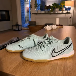 Nike fotbollsskor i mintgrönt - Säljer ett par mintgröna Nike fotbollsskor med svart swoosh och gummisula. Skorna har snörning, rund tå och är tillverkade i syntetmaterial och mesh för bra andningsförmåga. Perfekta för inomhusspel och har snygga detaljer på sulan. Använd 1-2 gånger nypris 1300 (hyfsat små i storleken)