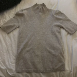 Beige polotröja med kort ärm - Säljer en stilren beige polotröja med korta ärmar och ribbade kanter. Tröjan har en hög krage och är gjord i mjukt material som känns skönt mot huden. Perfekt för lager-på-lager eller som den är till jeans eller kjol.