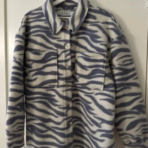 Zebramönstrad overshirt från Stay  - Overshirt i fleece från Stay med grått och vitt zebramönster. Skjortan har stora bröstfickor, klassisk krage och knäppning framtill. Perfekt för lager-på-lager och ger en cool vibe till din outfit.