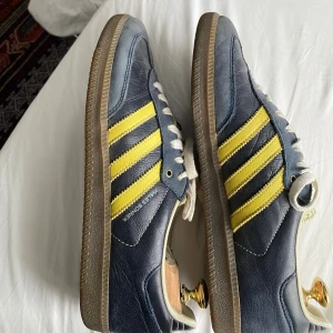 Adidas Wales Bonner Samba blå/gul - Säljer ett par Adidas Wales Bonner Samba sneakers i marinblått skinn med gula ränder och vit snörning. Klassisk gummisula och retrovibbar, perfekt för dig som gillar unika samarbeten och snygga detaljer. Ikonisk design med Wales Bonner-tryck på sidan.