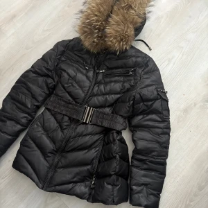 Svart pufferjacka med päls från Jollies - Svart pufferjacka från Jollies med fluffig brun päls på huvan, markerad midja med bälte och flera dragkedjor framtill. Jackan har lång ärm och quiltad design som ger en cool och snygg siluett. Perfekt för kalla dagar och vinterväder.