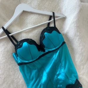 Turkos korsett med svarta spetsdetaljer - Snygg turkos korsett med svarta spetsdetaljer och justerbara axelband. Toppen har markerade sömmar, svarta band och spets längs byst och nederkant. Materialet är glansigt och känns lyxigt, med en croppad passform och hakknäppning bak. 