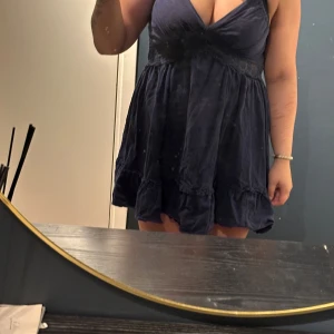 Mörkblå kort klänning med volang - Supersöt mörkblå kort klänning med djup v-ringning och justerbara smala axelband. Klänningen har volangdetaljer nertill och markerad midja för en feminin siluett. Perfekt för dig som gillar en luftig och lätt stil.