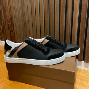 Burberry sneakers med klassiskt rutmönster - Snygga sneakers från Burberry med svart ovandel i skinn och mocka, vita sulor och märkets ikoniska beige och svarta rutmönster på sidorna. Skorna har snörning och en stilren, modern look som passar perfekt till streetwear eller en mer dressad outfit.