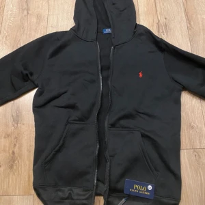 Svart hoodie från Polo Ralph Lauren - Svart hoodie från Polo Ralph Lauren med dragkedja och huva. Klassisk design med röd broderad logga på bröstet och två fickor framtill. Perfekt för en avslappnad stil och enkel att matcha med det mesta.