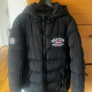 Svart pufferjacka från Polo Club - Svart pufferjacka från Polo Club med broderad logga och amerikansk flagga på bröstet. Jackan har huva med tryckt text, dragkedja framtill och två sidofickor. Perfekt för kalla dagar och har en schysst, sportig vibe.