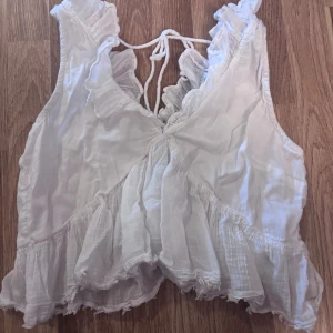 Vit volangig linnetopp med knyt - Supersöt vit linnetopp med volangdetaljer och knyt i ryggen. Toppen är ärmlös och har en luftig känsla med v-ringad skärning och romantiska volanger längs kanterna. Perfekt för varma dagar och en trendig look.