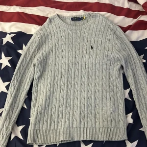 Ljusgrå stickad tröja från Polo Ralph Lauren - Säljer en ljusgrå stickad tröja från Polo Ralph Lauren med klassiskt kabelstickat mönster och broderad logga på bröstet. Tröjan har rund halsringning och långa ärmar, perfekt för lager-på-lager under kyliga dagar.