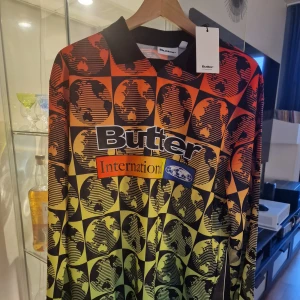 Butter International långärmad skateboardtröja - Snygg långärmad fotbollströja från Butter med grafiskt mönster av jordglobar i svart, rött, orange och gult. Tröjan har krage, tryckt logga och texten 'International' på bröstet. Tillverkad i ett lätt och glansigt material som känns sportigt och modernt.