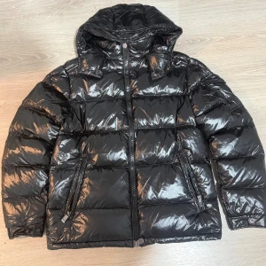 Moncler Maya - Snygg svart pufferjacka från Moncler med glansig finish och huva. Jackan har dragkedja framtill, två fickor med dragkedja och klassisk Moncler-logga på ärmen. Perfekt för kalla dagar när du vill hålla stilen och värmen.