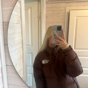 Brun pufferjacka från The North Face - Säljer en brun pufferjacka från The North Face i storlek S. Jackan har hög krage, dragkedja framtill och klassisk logga på bröstet. Den är vadderad och har en loose passform, perfekt för kalla dagar. Materialet är syntet och dun, vilket gör den riktigt varm och skön. Jackan har små defekter som även visas på en av bilderna!