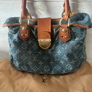 Louis Vuitton Monogram Denim Handväska - Snygg handväska från Louis Vuitton i blå denim med klassiskt monogrammönster och detaljer i brunt skinn. Väskan har robusta handtag, guldfärgade metalldetaljer och en unik låsdetalj framtill. Perfekt för dig som vill sticka ut med en ikonisk designeraccessoar. Väskan är köpt på soleplus och är vintage! Väskan saknar även två knappar och har defekter!