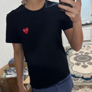 Svart Commes des Garçons Play t-shirt - Svart t-shirt från Commes des Garçons Play med klassisk rund hals och korta ärmar. På bröstet sitter det ikoniska röda hjärtat med ögon, broderad detalj. Materialet är mjuk bomull som känns skön mot huden. Perfekt för dig som gillar stilrena plagg med en twist.