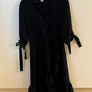 Svart omlottklänning från H&M Divided - Svart omlottklänning från H&M Divided med knytband vid ärmarna och volangdetalj längs nederkanten. Klänningen har v-ringning och trekvartsärmar, vilket ger en trendig och stilren look. Perfekt för dig som gillar minimalistisk stil med en twist.