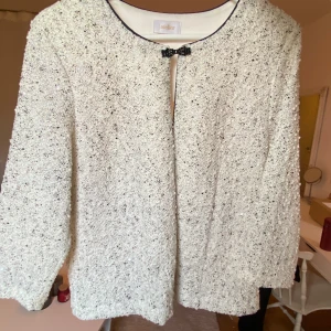 Vit cardigan jacka från Atelier - Säljer en vit cardigan jacka från Atelier med glittriga paljetter och svart rosett vid halsen. Jackan har rundad hals, lång ärm och svarta detaljer längs kanterna. Materialet är glittrigt och texturerat, vilket ger en lyxig känsla. Perfekt för dig som gillar att sticka ut. 