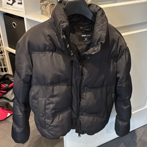 Svart pufferjacka  - Svart pufferjacka från F2 Sister i storlek XS. Jackan har hög krage, dragkedja och knappar framtill samt två fickor med lock. Den är vadderad och har en boxig, lite croppad passform som ger en cool vibe till din outfit. Jackan är knappt använd och är i nytt skick
