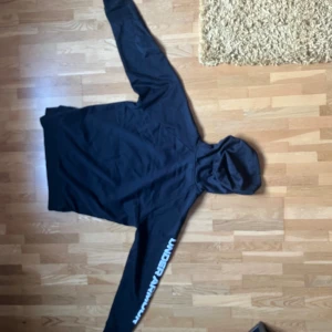 Svart hoodie från Under Armour - Svart hoodie från Under Armour med logga på ena ärmen och liten logga framtill. Klassisk huva med snörning och stor magficka. Perfekt för chill eller träning, riktigt skön passform och stilren look.