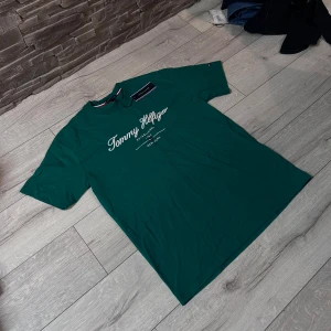 Grön Tommy Hilfiger t-shirt med tryck - Snygg grön t-shirt från Tommy Hilfiger med vit broderad logga och text på bröstet. Klassisk rund hals och korta ärmar. Tillverkad i mjuk bomull för skön känsla. Perfekt för dig som gillar stilrena och ikoniska plagg med tydlig branding.