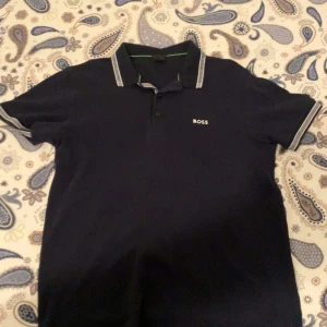 Marinblå pikétröja från BOSS - Snygg marinblå pikétröja från BOSS med vit logga på bröstet. Tröjan har klassisk krage, korta ärmar och vita detaljer på krage och ärmslut. Perfekt för dig som gillar stilren design och vill ha en tröja med lite edge.