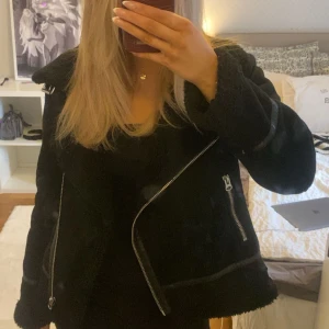 Svart teddyjacka från Zara med dragkedjor - Säljer en svart teddyjacka från Zara i storlek M. Jackan har grov krage, flera silvriga dragkedjor och snygga detaljer i skinnimitation. Fodret är fluffigt och jackan har en loose passform med långärmade ärmar. Perfekt för kalla dagar och riktigt trendig look.