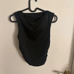 Svart ärmlös topp med rynkade sidor - En svart ärmlös topp med snygg rynkdetalj längs sidorna. Toppen har en enkel och stilren design med rundad halsringning och är tillverkad i ett mjukt, stretchigt material som sitter bekvämt mot kroppen.