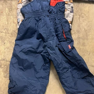 Mörkblå täckbyxor med hängslen - Mörkblå täckbyxor med elastisk midja och justerbara hängslen. Byxorna har en röd detalj vid fickan och är tillverkade i ett slitstarkt, vattenavvisande material som passar perfekt för kalla dagar utomhus.