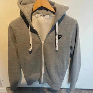 Grå hoodie från Comme des Garçons Play - Grå hoodie från Comme des Garçons Play med dragkedja, vit snörning och huva. På bröstet sitter det ikoniska svarta hjärta med ögon. Tröjan har mjukt fleecefoder och två fickor framtill. Perfekt för chill dagar och streetstyle.