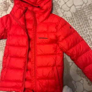 Röd dunjacka Peak Performance XS - Riktigt snygg röd dunjacka från Peak Performance i storlek XS. Jackan har huva, dragkedja och är quiltad för extra värme. Perfekt för kyliga dagar och har en sportig vibe. Logga på bröstet och ärmen. Fylld med certifierat dun. Helt ny säljer då den va för liten, taggarna sitter kvar 