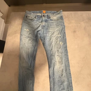 Boss Orange ljusblå jeans - Boss Orange ljusblå jeans med slitna detaljer och klassisk femficksdesign. Jeansen har raka ben och är tillverkade i mjukt bomullsjeansmaterial. Snyggt orange Boss-märke i midjan och läderpatch baktill. Perfekt för dig som gillar en avslappnad och trendig look.