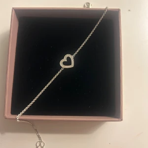 Thomas Sabo armband med hjärta i silver - Superfint armband från Thomas Sabo i äkta silver med en öppen hjärtformad detalj i mitten, dekorerad med små gnistrande stenar. Tunn kedja och smidig karbinhake. Perfekt accessoar för att lyfta din stil. Nypris: 949 kr 