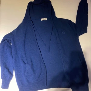 Marinblå hoodie i merinoull & kashmir - Mysig marinblå hoodie från AW i lyxig mix av 90% merinoull och 10% kashmir. Tröjan har huva med snörning, långa ärmar och två fickor framtill. Perfekt för dig som gillar en stilren och bekväm look med premiumkänsla.