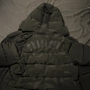 Svart pufferjacka Trapstar XS - Svart pufferjacka från Trapstar i storlek XS. Jackan har en stor huva, broderad Trapstar-logga på ryggen och dragkedja framtill. Tillverkad i 100% polyester med ett clean och stilrent utseende, perfekt för kalla dagar.