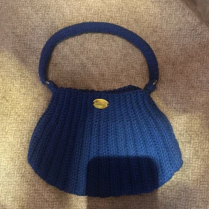 Blå stickad handväska med guldmärke - Superfin blå handväska i stickat material med bred axelrem och ett litet guldfärgat märke framtill där det står 'Handmade'. Väskan har en mjuk, ribbad struktur och en unik, rundad form som sticker ut.