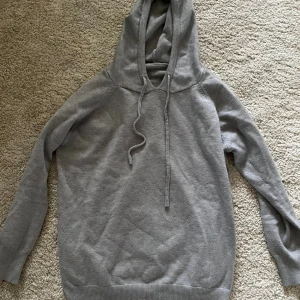 Grå stickad hoodie med snörning - Mjuk och enkel grå hoodie med huva och snörning framtill. Stickad känsla och ribbade muddar vid ärmslut och nederkant. Perfekt för chill dagar och när du vill ha något bekvämt och stilrent.