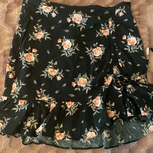 Gullig blommig kjol från hm  - Svart kjol med blommor på. Den kommer från hm och är i storlek s. Kjolen har volanger nere och har en knyt detalj där man kan bestämma hur hög slitsen ska vara