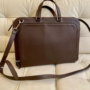 Prada portfölj, Saffiano Travel - Snygg, klassisk mörkbrun portfölj i kalvskinn från Prada. Perfekt för laptop och dokument. I mycket gott skick, så gott som ny, inga skador eller repor.  Modell Saffiano Travel, färg Caffé.  Inköpt på Pradabutiken i Stockholm. Äkthetsintyg medföljer. Nypris ca 16.000:-